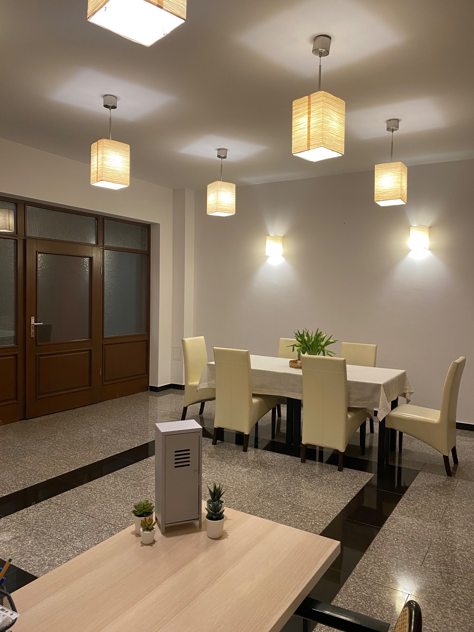 Sala konferencyjna z dużym stołem, sześcioma krzesłami, dekoracyjnymi lampami sufitowymi i ściennymi oraz roślinami doniczkowymi na stole i małej szafce.