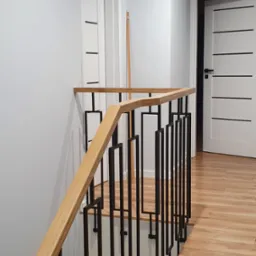Nowoczesna balustrada schodowa z jasnego drewna i czarnych, prostokątnych metalowych elementów, widok z dołu schodów, w tle białe drzwi z czarnymi akcentami i podłoga imitująca drewno.