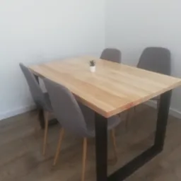 Jasny, prostokątny blat drewniany na czarnych, metalowych nogach, otoczony czterema szarymi krzesłami na drewnianych nogach, z małą rośliną doniczkową na środku blatu.