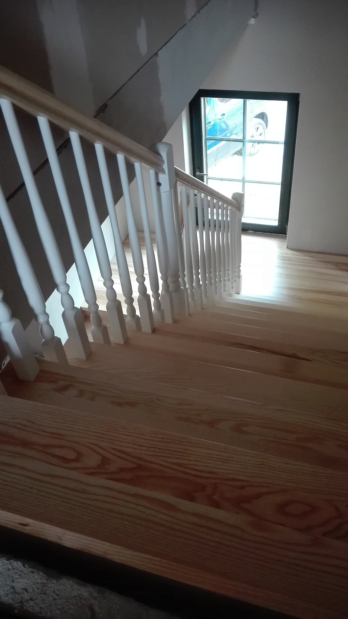 Jasne, drewniane schody z białą balustradą w trakcie montażu, widoczne surowe ściany i okno z widokiem na samochód.