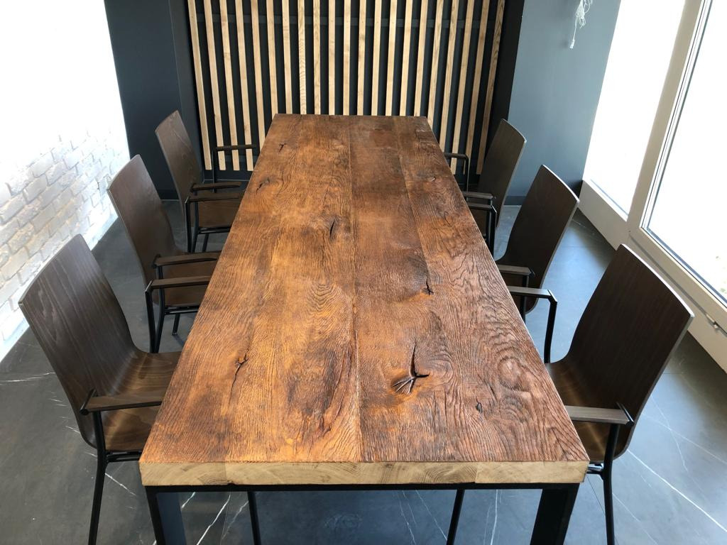 Długi, rustykalny blat drewniany z widocznymi sękami i naturalnym usłojeniem, osadzony na czarnej, metalowej ramie stołu, otoczony sześcioma krzesłami z drewnianymi oparciami i metalowymi nogami...