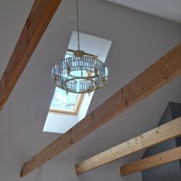 Maksbud Maksym Rosenko - Kryształowy żyrandol zawieszony na łańcuchu, widoczny na tle okna dachowego, drewniane belki stropowe w jasnym pomieszczeniu z szarą ścianą.