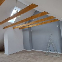 Maksbud Maksym Rosenko - Wykończone poddasze ze skośnymi ścianami, drewnianymi belkami stropowymi, oknem dachowym i szarą ścianą. Na podłodze panele, widoczna drabina malarska.