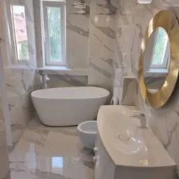 Wykończona łazienka w marmurowej stylistyce z wolnostojącą wanną, bidetem, umywalką na szafce i okrągłym lustrem w złotej ramie, oświetlona nowoczesnymi lampami sufitowymi.