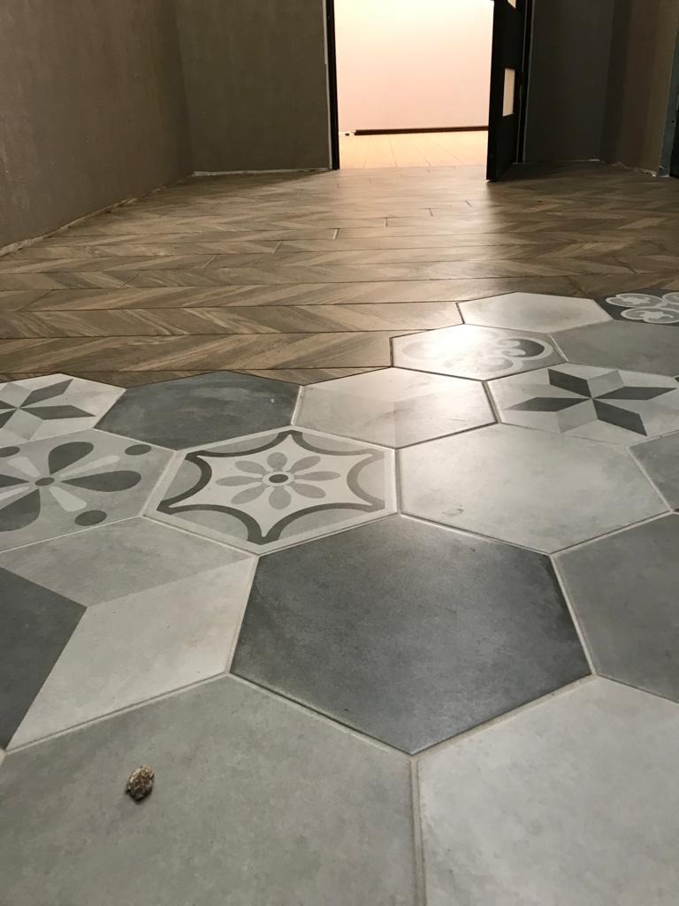 Połączenie podłogi z drewnopodobnych paneli ułożonych w jodełkę z heksagonalnymi płytkami ceramicznymi w odcieniach szarości i bieli ze wzorami kwiatowymi, widok na otwarte drzwi do innego...