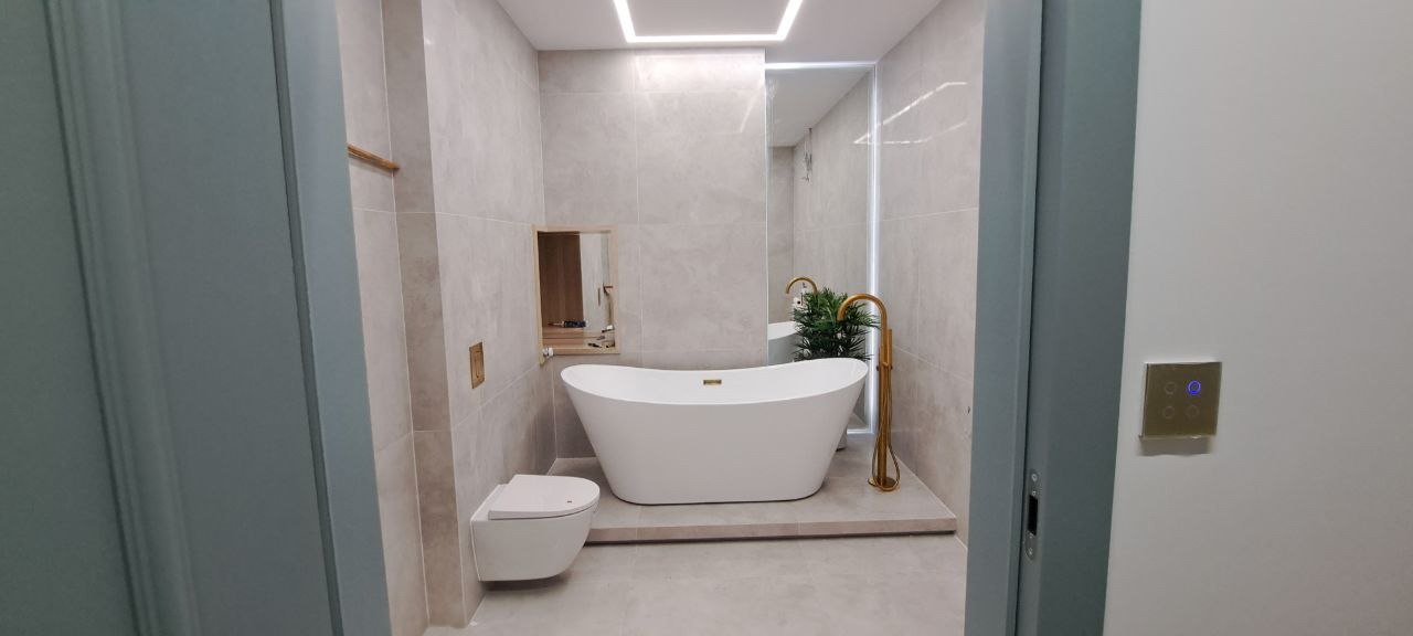 Nowoczesna łazienka z wolnostojącą białą wanną, złotą armaturą i podwieszaną toaletą, wykończona w jasnych płytkach z subtelnym oświetleniem LED.