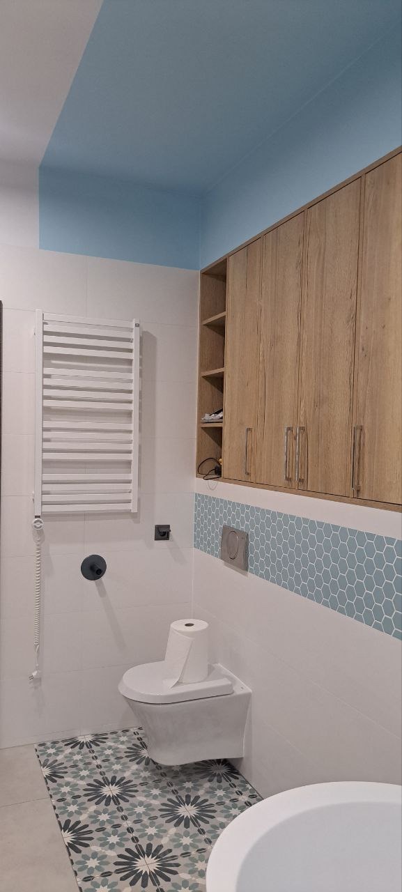 Nowoczesna łazienka z wiszącą toaletą, grzejnikiem drabinkowym, drewnianymi szafkami i dekoracyjnymi płytkami heksagonalnymi oraz wzorzystą podłogą.