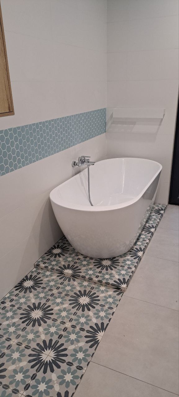 Biała, wolnostojąca wanna na tle łazienki z dekoracyjnymi, wzorzystymi płytkami podłogowymi i pasem mozaiki heksagonalnej na ścianie.