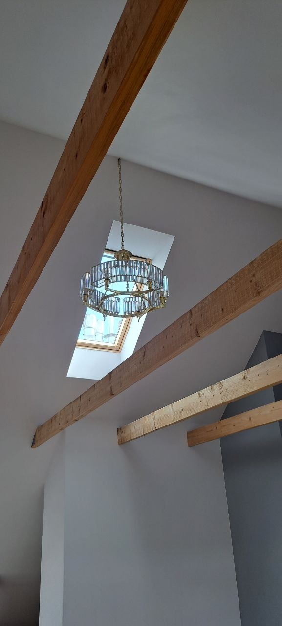 Kryształowy żyrandol zawieszony na łańcuchu, widoczny na tle okna dachowego, drewniane belki stropowe w jasnym pomieszczeniu z szarą ścianą.