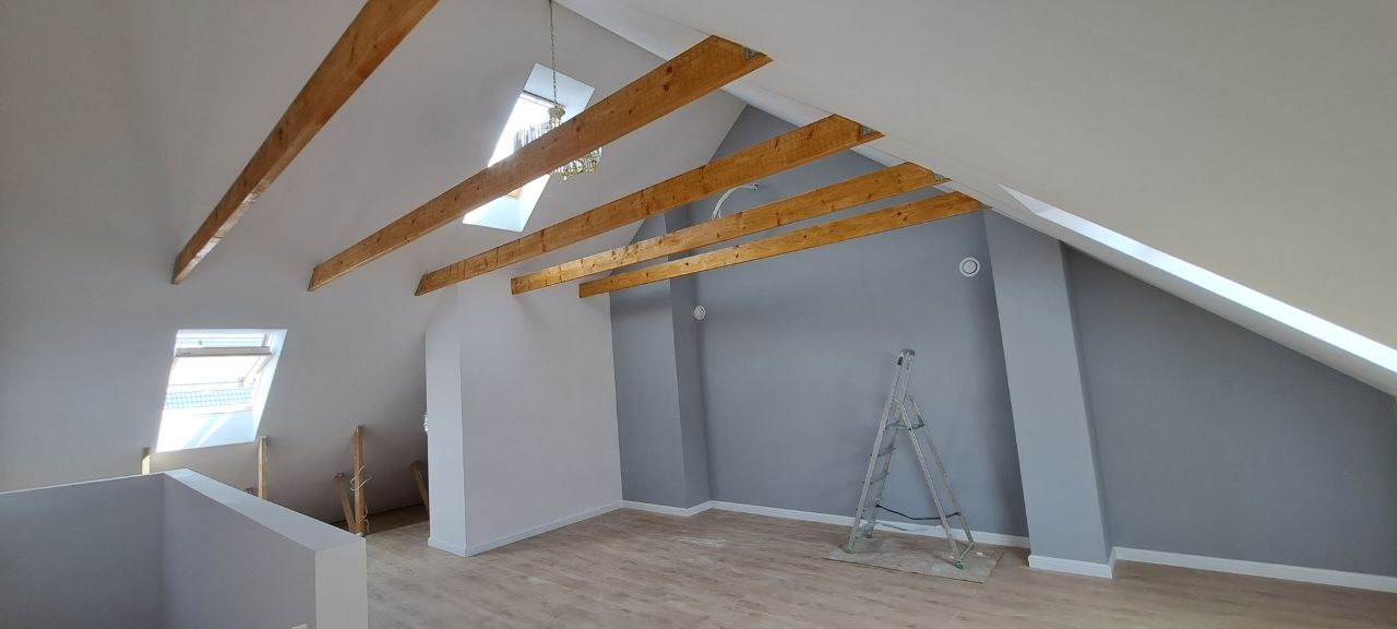 Wykończone poddasze ze skośnymi ścianami, drewnianymi belkami stropowymi, oknem dachowym i szarą ścianą. Na podłodze panele, widoczna drabina malarska.