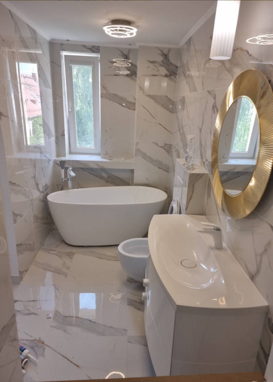 Wykończona łazienka w marmurowej stylistyce z wolnostojącą wanną, bidetem, umywalką na szafce i okrągłym lustrem w złotej ramie, oświetlona nowoczesnymi lampami sufitowymi.