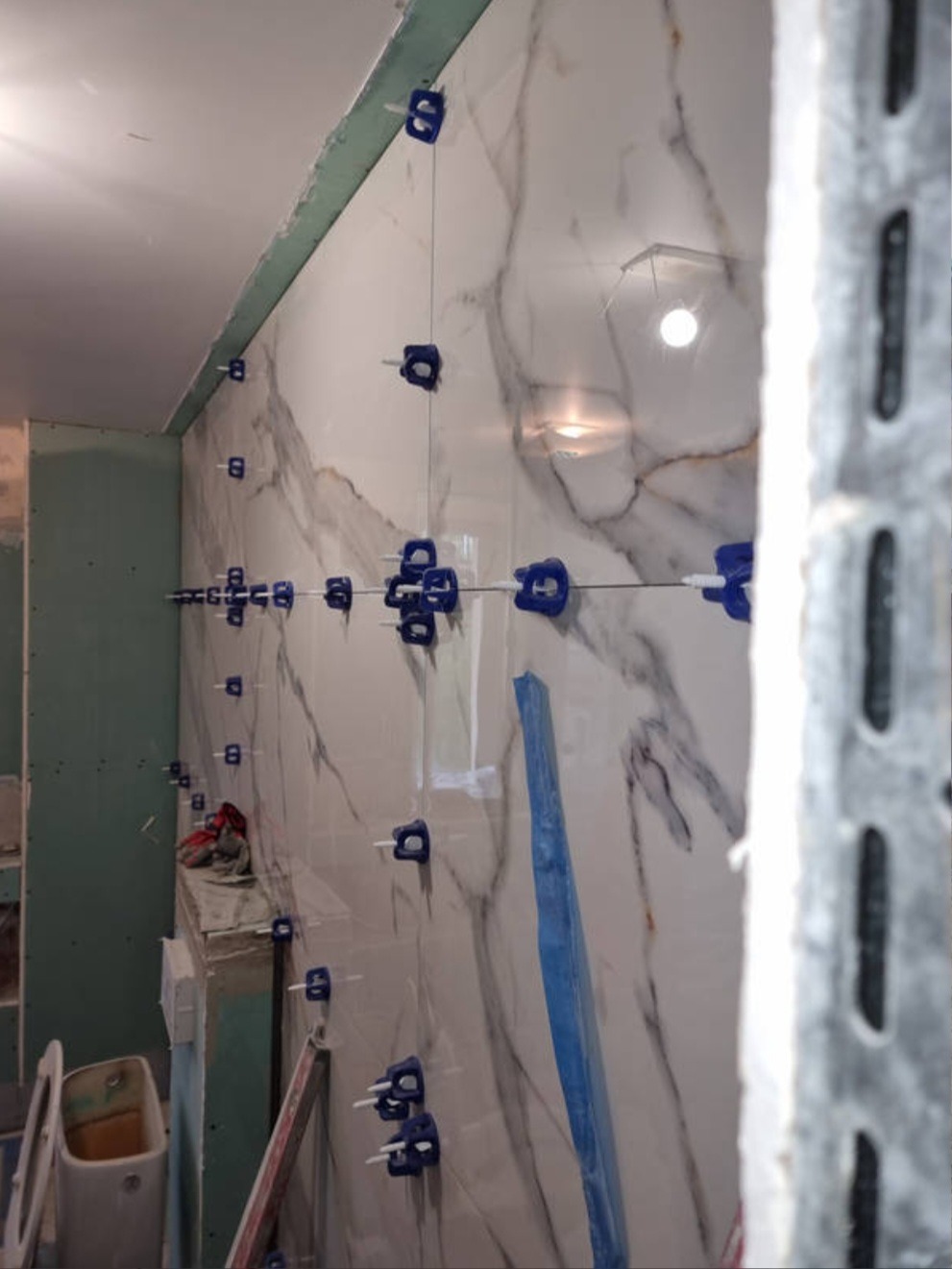 Ściana wyłożona białymi płytkami imitującymi marmur, z widocznymi niebieskimi klipsami poziomującymi i sznurkiem, w trakcie prac wykończeniowych w łazience, widoczny fragment toalety i płyta karton...