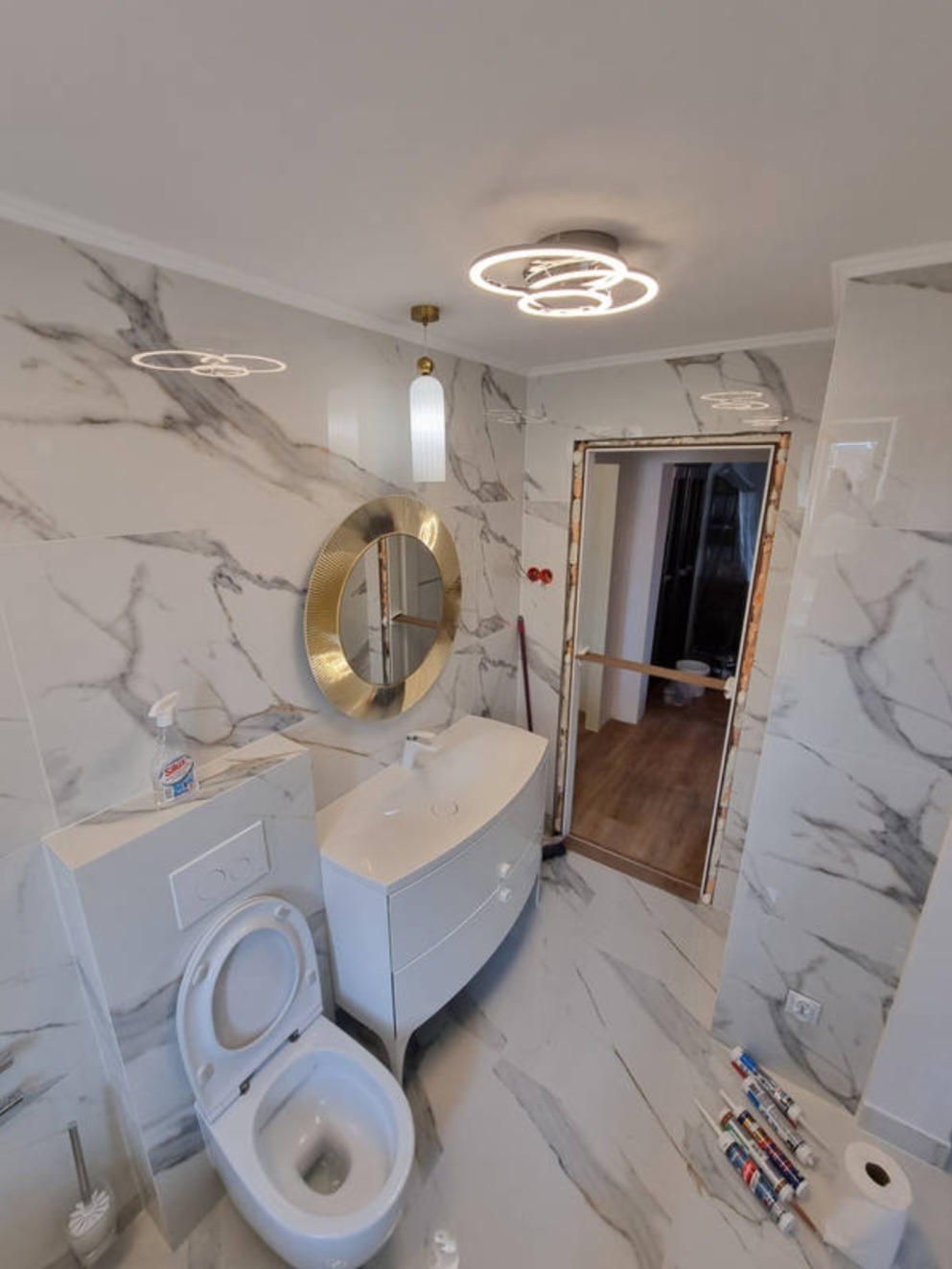 Nowoczesna łazienka z marmurowymi płytkami, okrągłym lustrem w złotej ramie, białą szafką pod umywalką i toaletą, widoczne tuby z silikonem na podłodze, sufit z ozdobnym oświetleniem.