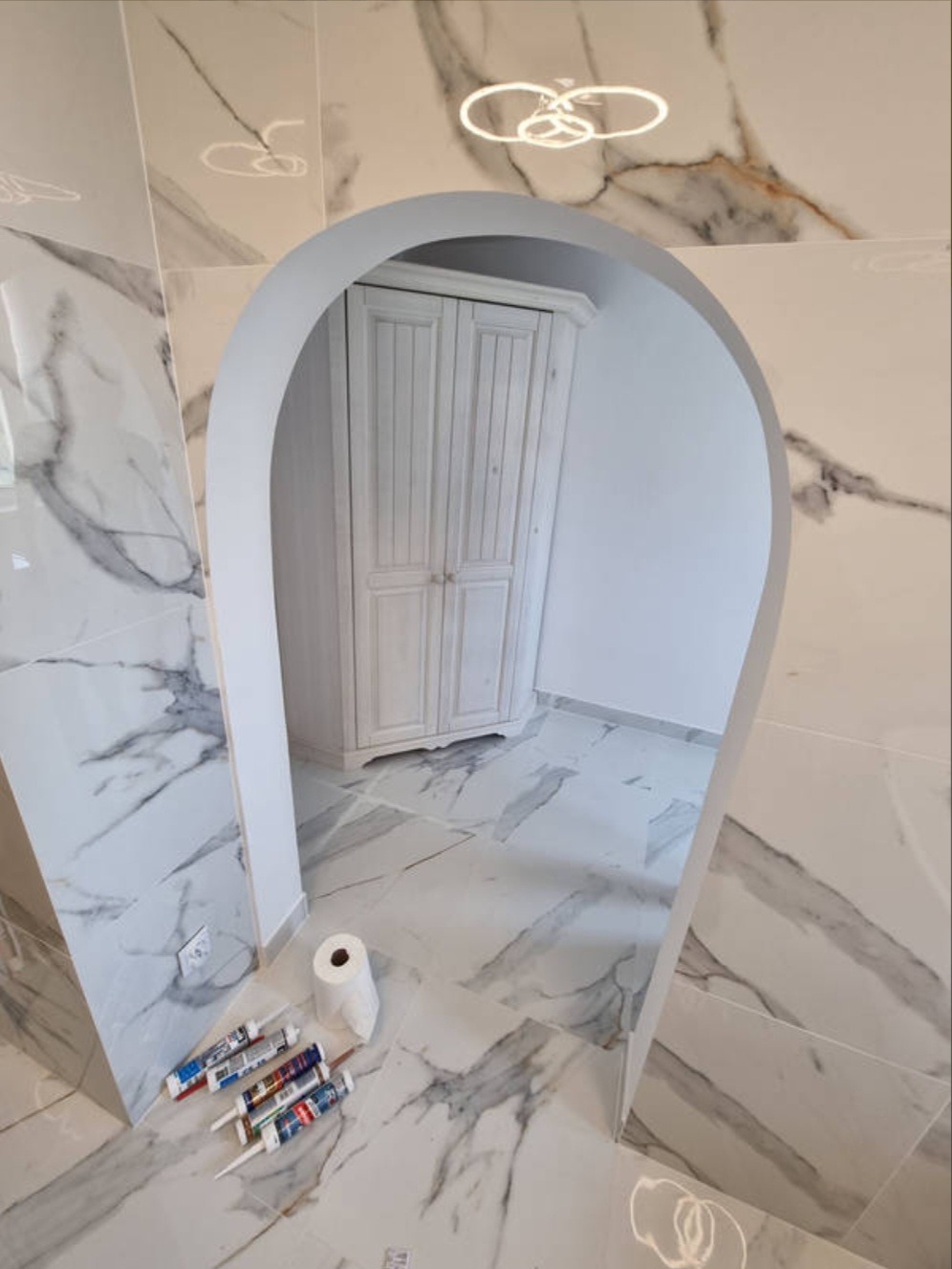 Łazienka z marmurowymi płytkami i łukowym wejściem, widoczne tubki silikonu i rolka papieru toaletowego na podłodze, w głębi biała szafka.