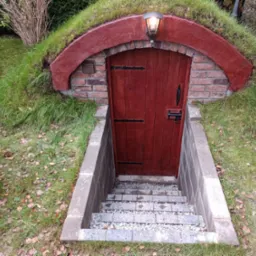 Wejście do ziemianki z drewnianymi drzwiami, ceglanym obramowaniem i schodami z kostki brukowej, porośnięte trawą i oświetlone lampą.