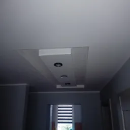 Szare wnętrze z podwieszanym sufitem z płyt kartonowo-gipsowych z trzema wpuszczonymi punktami świetlnymi, widok na okno z żaluzjami.