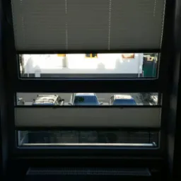 Dwuczęściowe, jasne osłony okienne plisowane na oknie z widokiem na podwórze z zaparkowanymi samochodami i domem z kominem w tle.