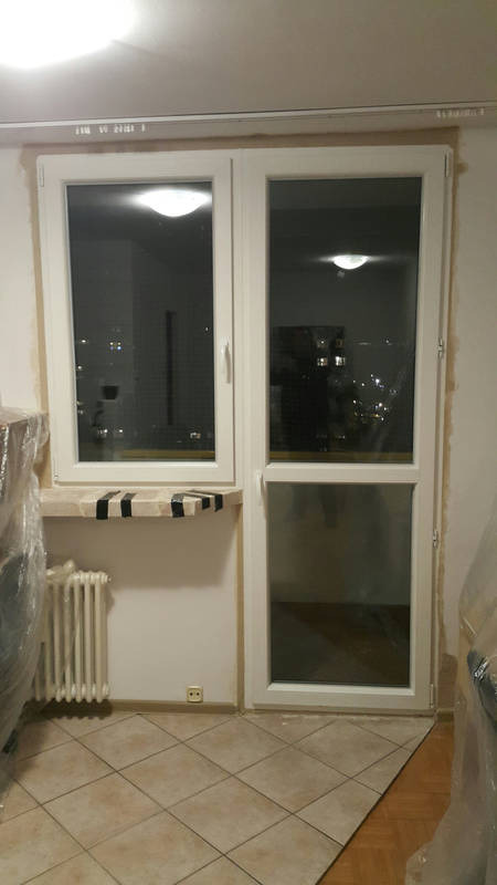 Białe okno balkonowe z pojedynczym skrzydłem i częścią stałą, widok na nocne miasto, zabezpieczony parapet taśmą malarską, grzejnik żeberkowy.