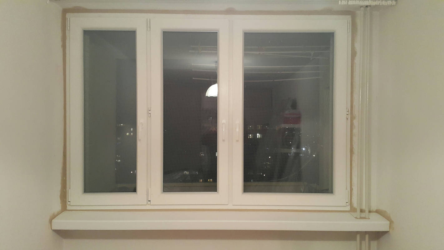 Trzyczęściowe białe okno z widokiem na nocne miasto, widoczne ślady prac wykończeniowych na ścianie i ramie okiennej.