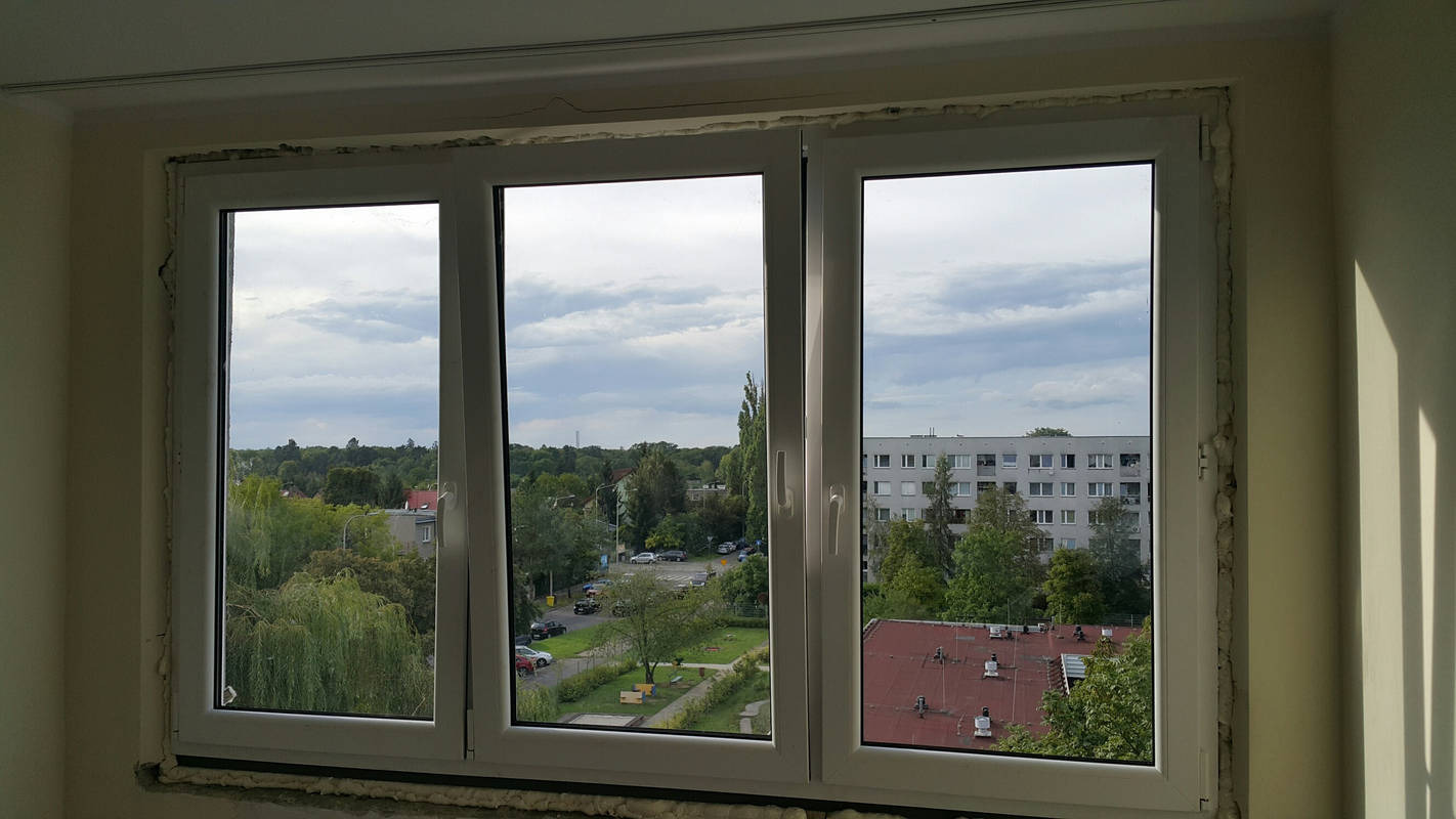Trzyszybowe okno z białymi ramami, widok na osiedle z zielenią, pianka montażowa wokół ramy okiennej, ślady zabrudzeń na ścianie nad oknem.