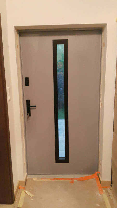 Szare drzwi wejściowe z pionowym, prostokątnym przeszkleniem, widoczna czarna klamka i zamek, zabezpieczenia podłogi taśmą malarską podczas montażu.