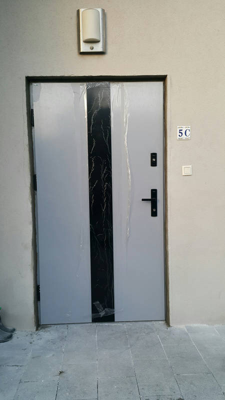 Szare drzwi wejściowe z czarnym, pionowym panelem dekoracyjnym, częściowo owinięte folią ochronną, zamontowane w jasnej ścianie budynku z numerem 5C i lampą zewnętrzną.