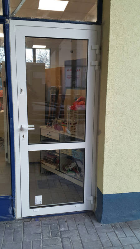 Białe drzwi wejściowe z PCV z dwoma przeszkleniami, widok z zewnątrz, z widocznym wnętrzem sklepu lub biura.