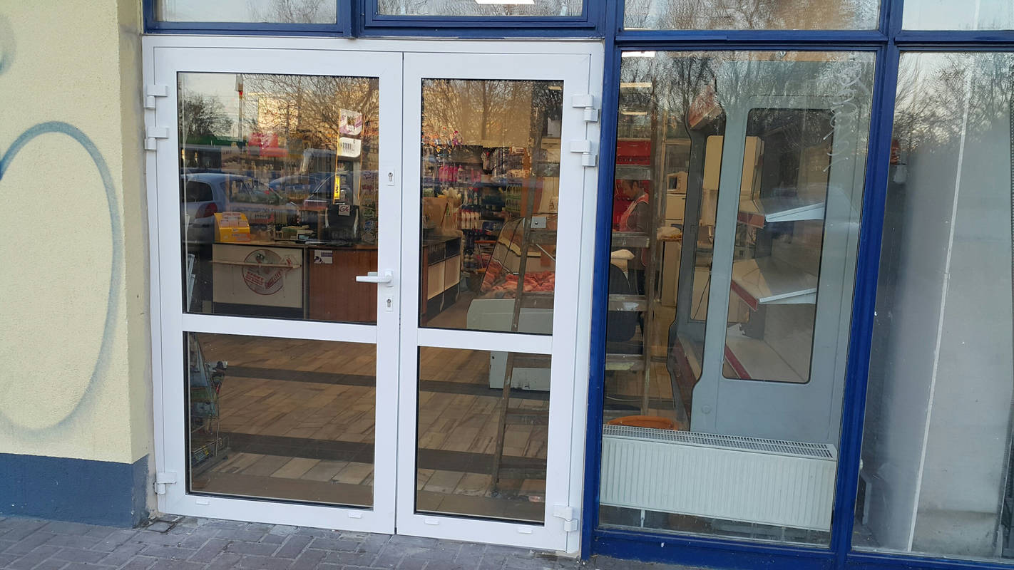 Białe, dwuskrzydłowe drzwi wejściowe do sklepu, widok z zewnątrz, z refleksami wnętrza sklepu i ulicy.