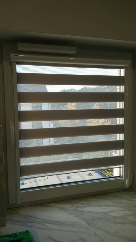 Beżowo-biała roleta dzień noc zamontowana w oknie PCV, widok na drzewa za oknem, marmurowy parapet.