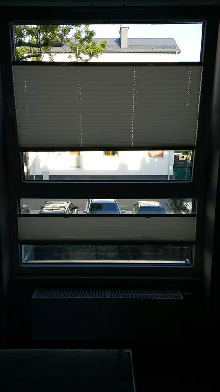 Dwuczęściowe, jasne osłony okienne plisowane na oknie z widokiem na podwórze z zaparkowanymi samochodami i domem z kominem w tle.