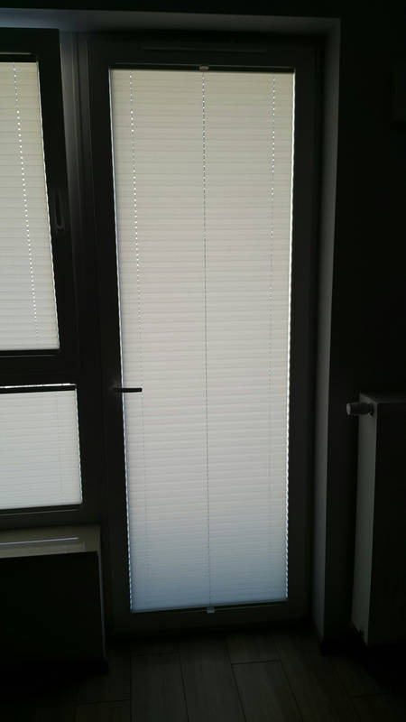 Zamknięte białe plisy okienne w oknie PCV, widoczne poziome lamele i mechanizm sterujący, ujęcie z wnętrza pomieszczenia.