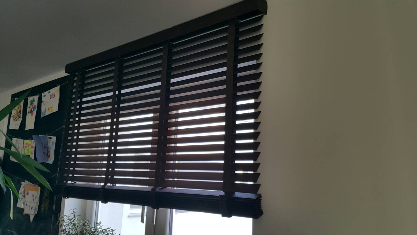 Ciemnobrązowe żaluzje poziome drewniane zamontowane w oknie, częściowo zasłonięte, z widocznymi rysunkami dzieci na tablicy w tle i roślinami doniczkowymi na parapecie.