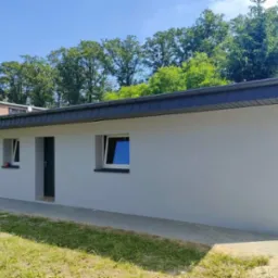 Nowoczesny, pomalowany na szaro budynek z grafitowym dachem i rynnami, widok z zewnątrz w słoneczny dzień, otoczony zielenią.