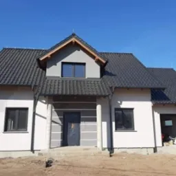 Dom jednorodzinny w trakcie budowy, z grafitową dachówką karpiówką, widoczne rynny, elewacja w stanie surowym, niezakończone prace wykończeniowe wokół budynku.