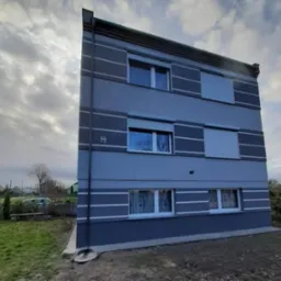 Trzypiętrowy budynek po renowacji elewacji w odcieniach szarości z białymi akcentami, widoczne okna z roletami i firankami, na tle pochmurnego nieba.