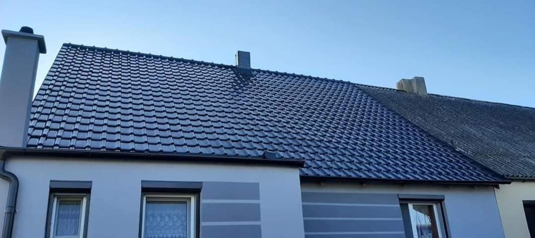 Nowy, błyszczący dach z ciemnej dachówki ceramicznej na tle błękitnego nieba, sąsiadujący ze starym, pokrytym mchem dachem z eternitu.