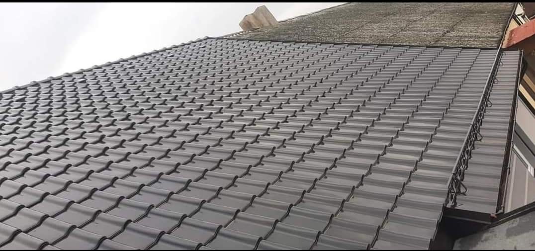 Nowy, grafitowy dach z dachówki ceramicznej, widoczny z dołu pod kątem, z fragmentem starego dachu z papy w tle. Zauważalny system rynnowy wzdłuż krawędzi dachu.