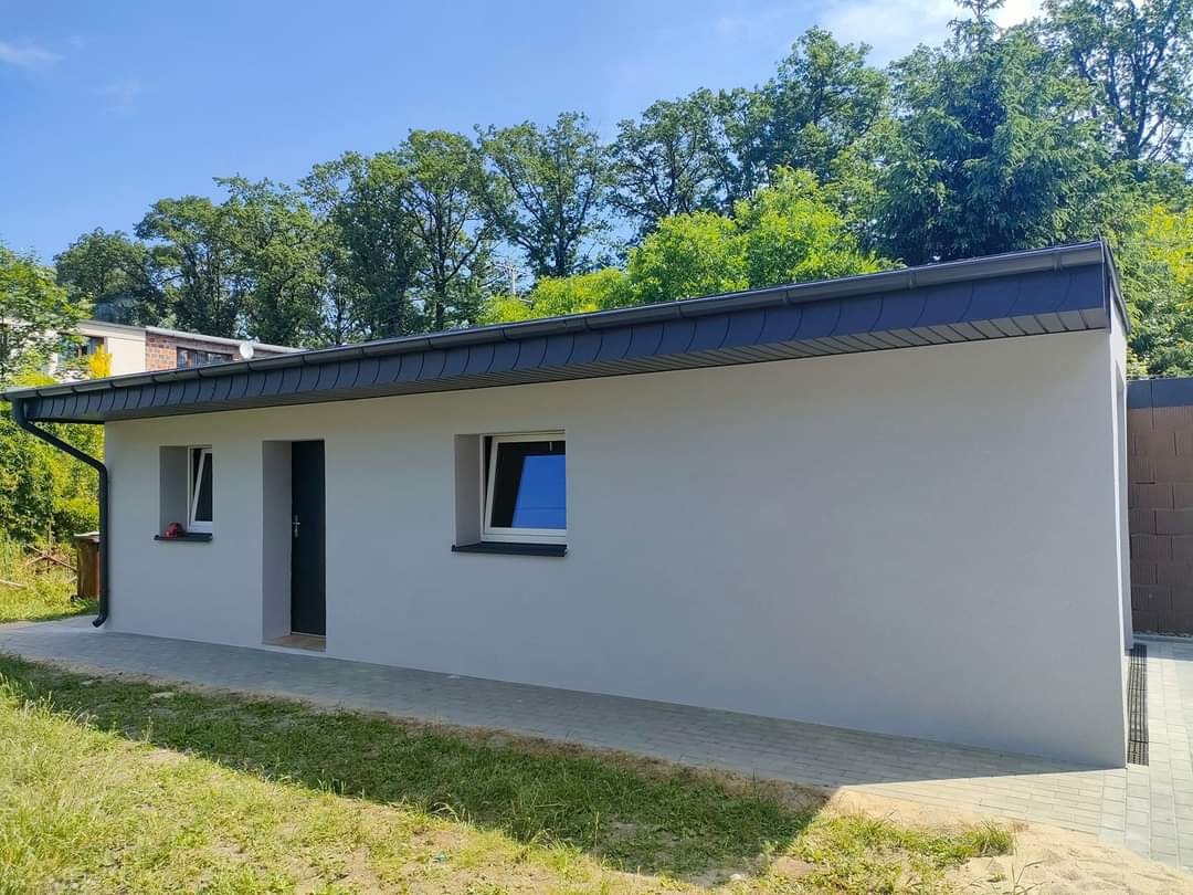 Nowoczesny, pomalowany na szaro budynek z grafitowym dachem i rynnami, widok z zewnątrz w słoneczny dzień, otoczony zielenią.