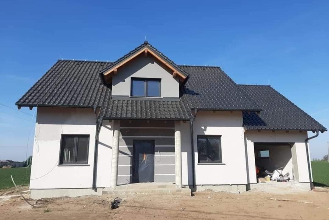 Dom jednorodzinny w trakcie budowy, z grafitową dachówką karpiówką, widoczne rynny, elewacja w stanie surowym, niezakończone prace wykończeniowe wokół budynku.