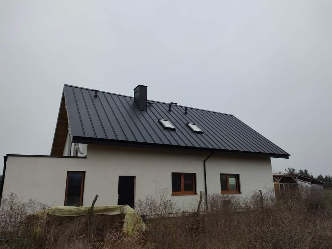 Dom jednorodzinny z nowym, ciemnoszarym dachem z blachy na rąbek stojący, widoczne okna dachowe i komin. Elewacja budynku w jasnym kolorze, teren wokół częściowo zagospodarowany.