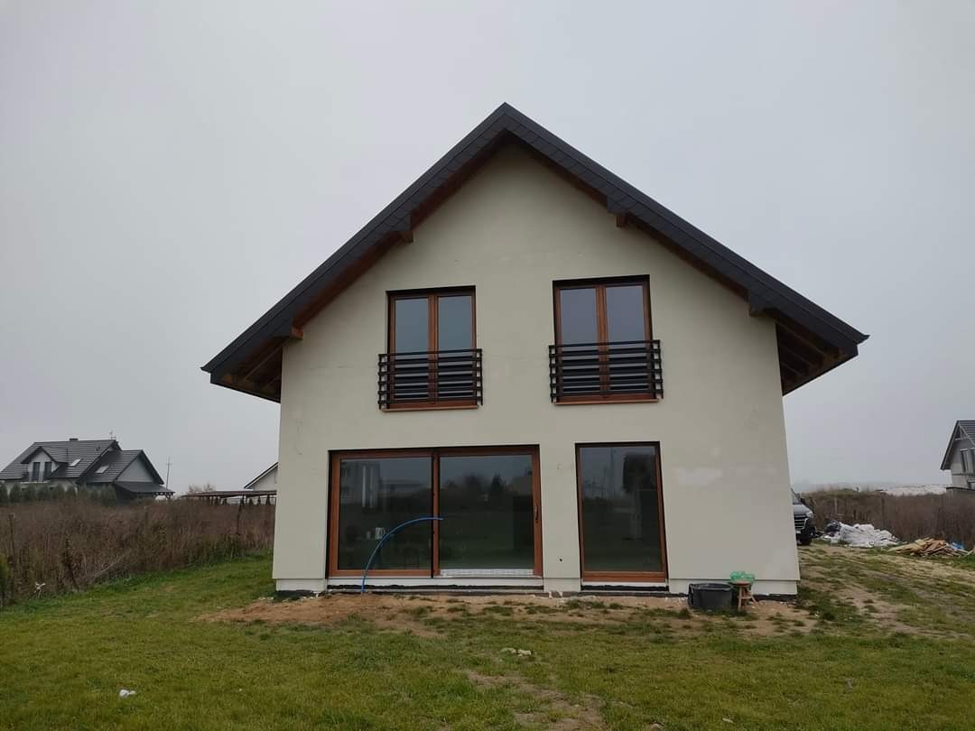 Nowoczesny dom jednorodzinny z jasną elewacją, ciemnym dachem i drewnianymi oknami, widoczne ślady po pracach elewacyjnych, trawa i niebo pochmurne.
