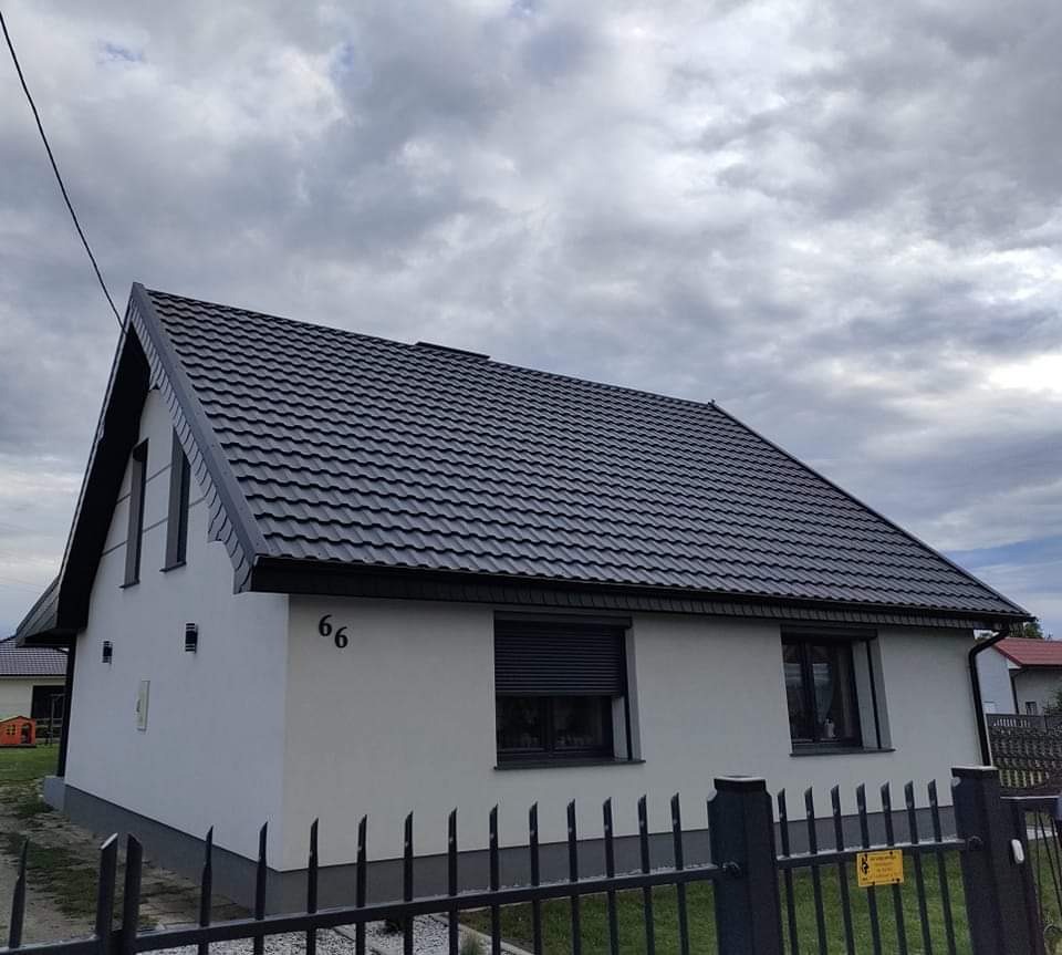 Dom jednorodzinny z nowym, ciemnoszarym dachem z blachodachówki modułowej, pomalowaną elewacją na biało i numerem '66' na ścianie, widok zza czarnego metalowego ogrodzenia.