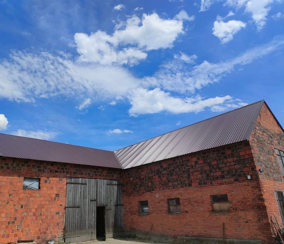 Budynek gospodarczy z czerwonej cegły z nowym, brązowym dachem z blachy trapezowej na tle błękitnego nieba z białymi chmurami.