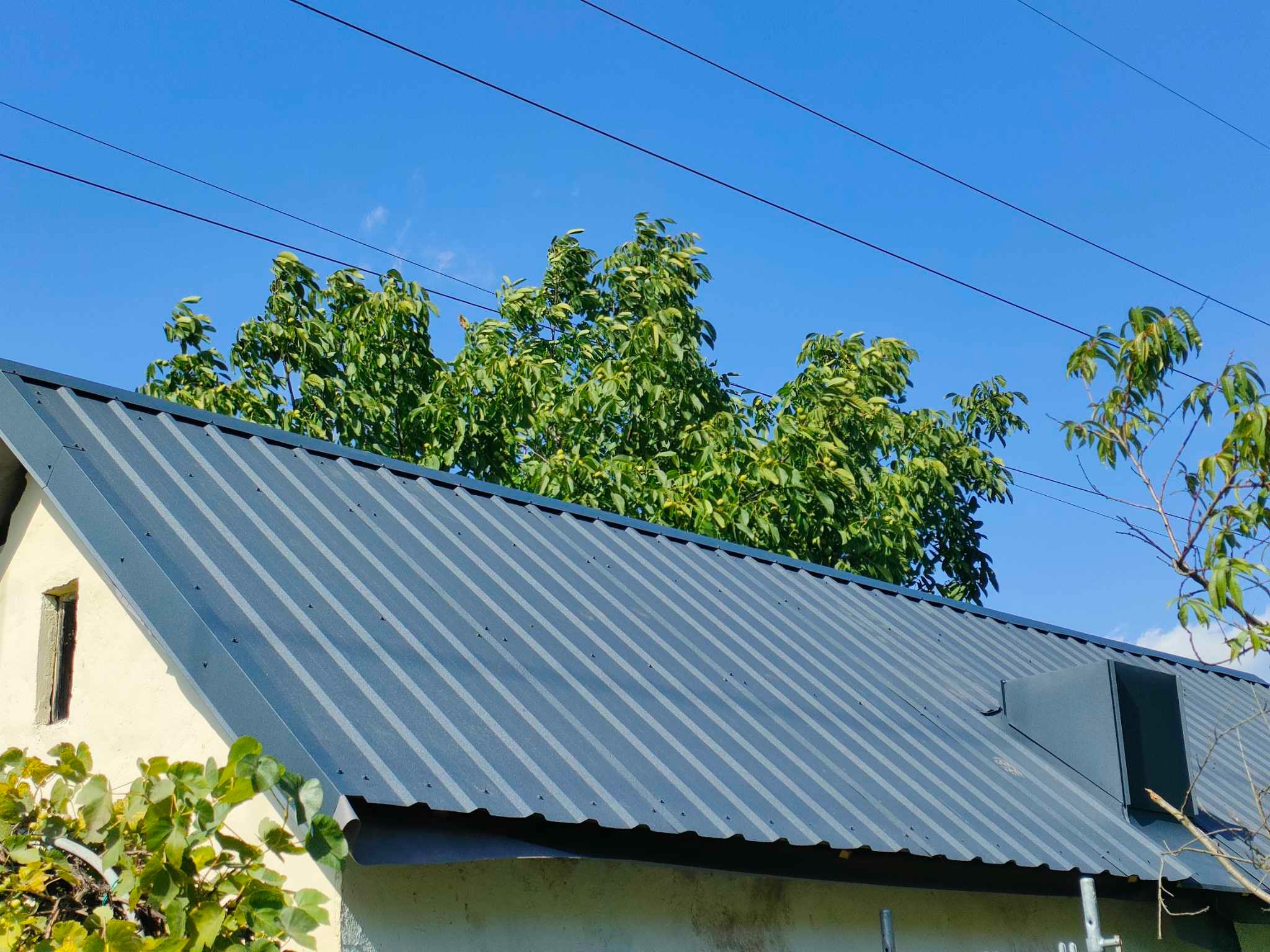 Szary dach z blachy trapezowej na budynku jednorodzinnym, widok z dołu, w tle błękitne niebo i korony drzew.