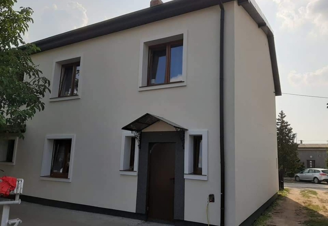 Dwupiętrowy dom z jasną elewacją, ciemnymi ramami okiennymi i drzwiami wejściowymi z ozdobnym daszkiem, widok z zewnątrz po renowacji.