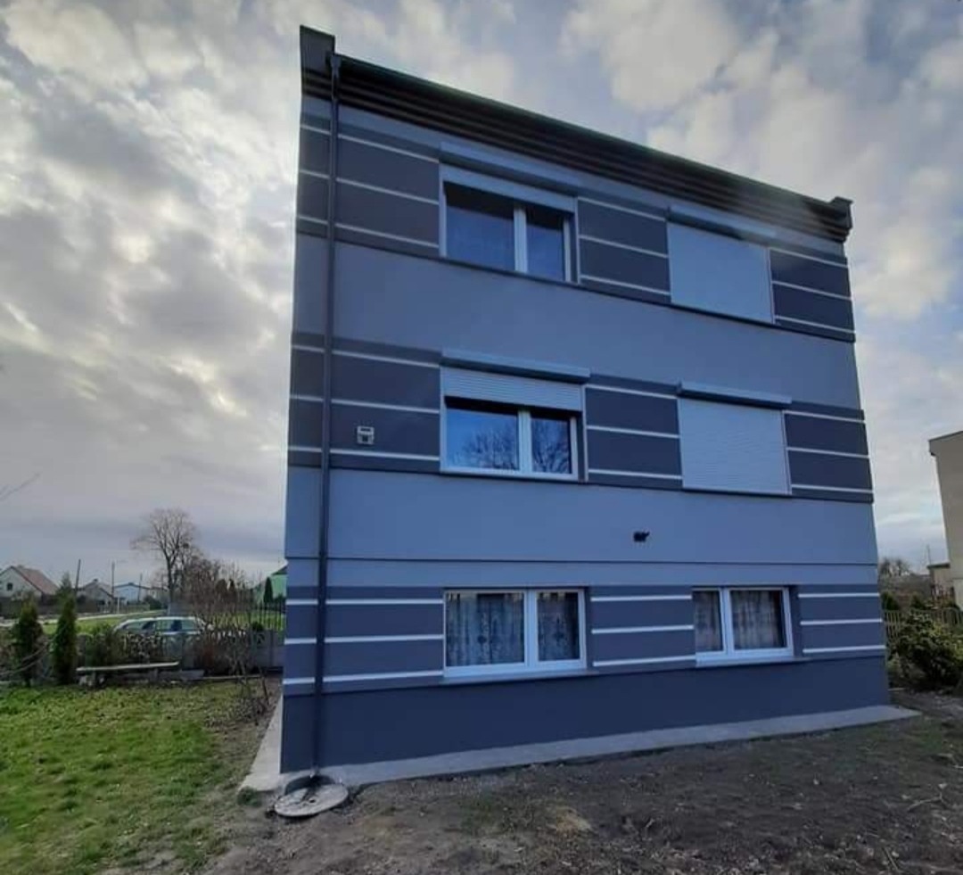 Trzypiętrowy budynek po renowacji elewacji w odcieniach szarości z białymi akcentami, widoczne okna z roletami i firankami, na tle pochmurnego nieba.