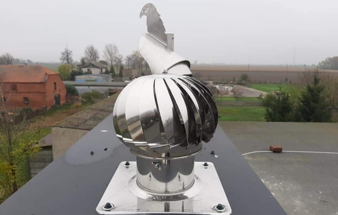 Błyszcząca, metalowa turbina dachowa z motywem orła na szczycie, zamontowana na ciemnym dachu z widokiem na wiejski krajobraz w tle.