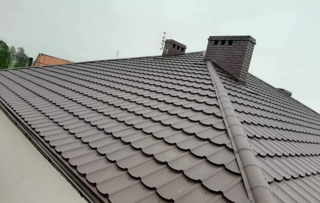 Nowy, brązowy dach z blachodachówki modułowej z kominem z cegły klinkierowej i anteną telewizyjną, widziany pod kątem od dołu.