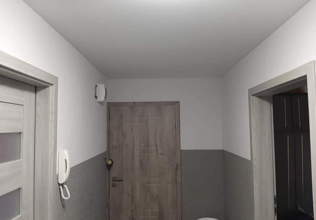 Wnętrze korytarza po remoncie, z pomalowanymi ścianami na biało i szaro, widoczne drzwi wejściowe z drewnopodobną okleiną, drzwi wewnętrzne z szarą ramą oraz domofon na ścianie.