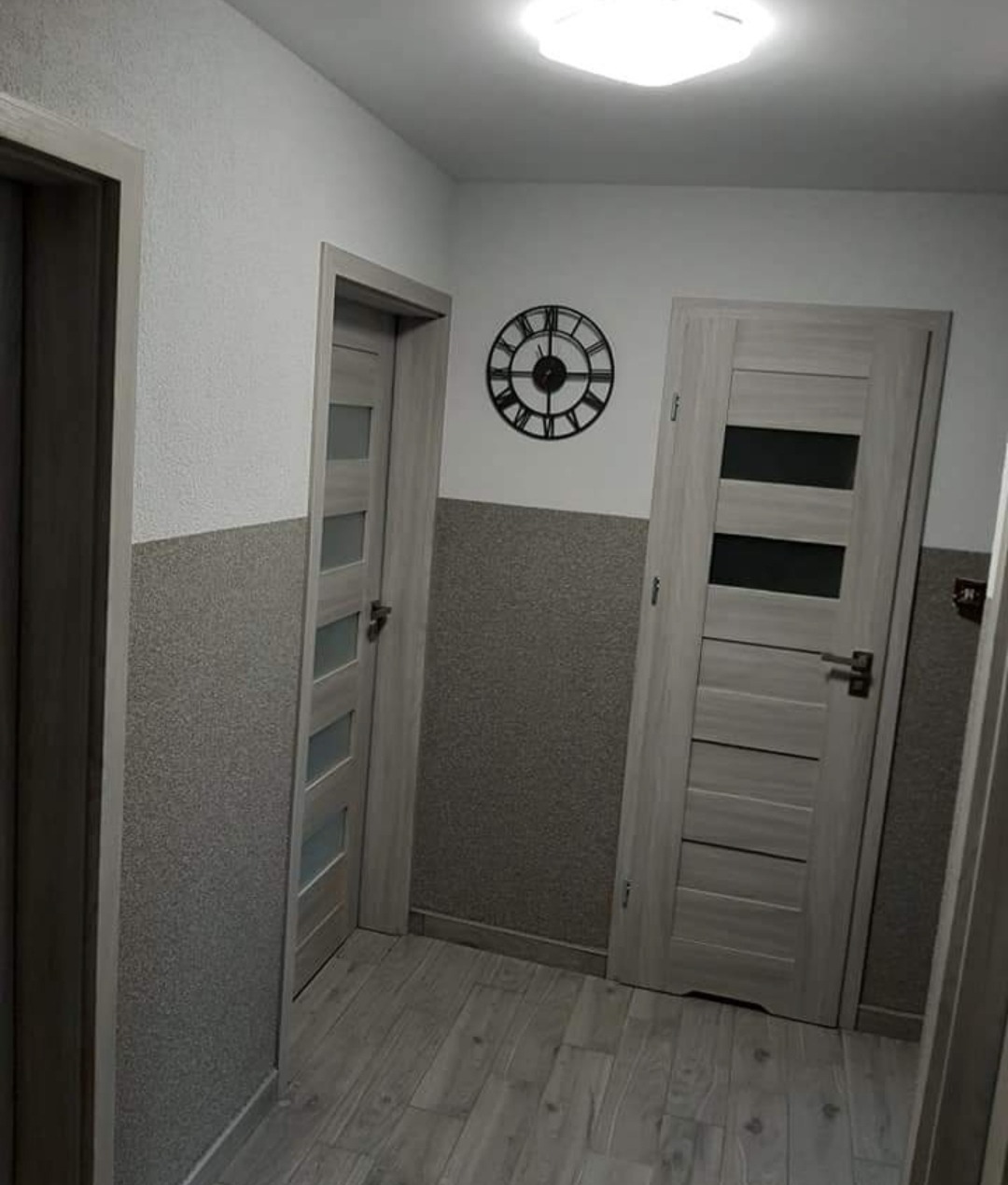 Korytarz z dwoma szarymi drzwiami wewnętrznymi z przeszkleniami, szarą boazerią i zegarem ściennym w stylu industrialnym.